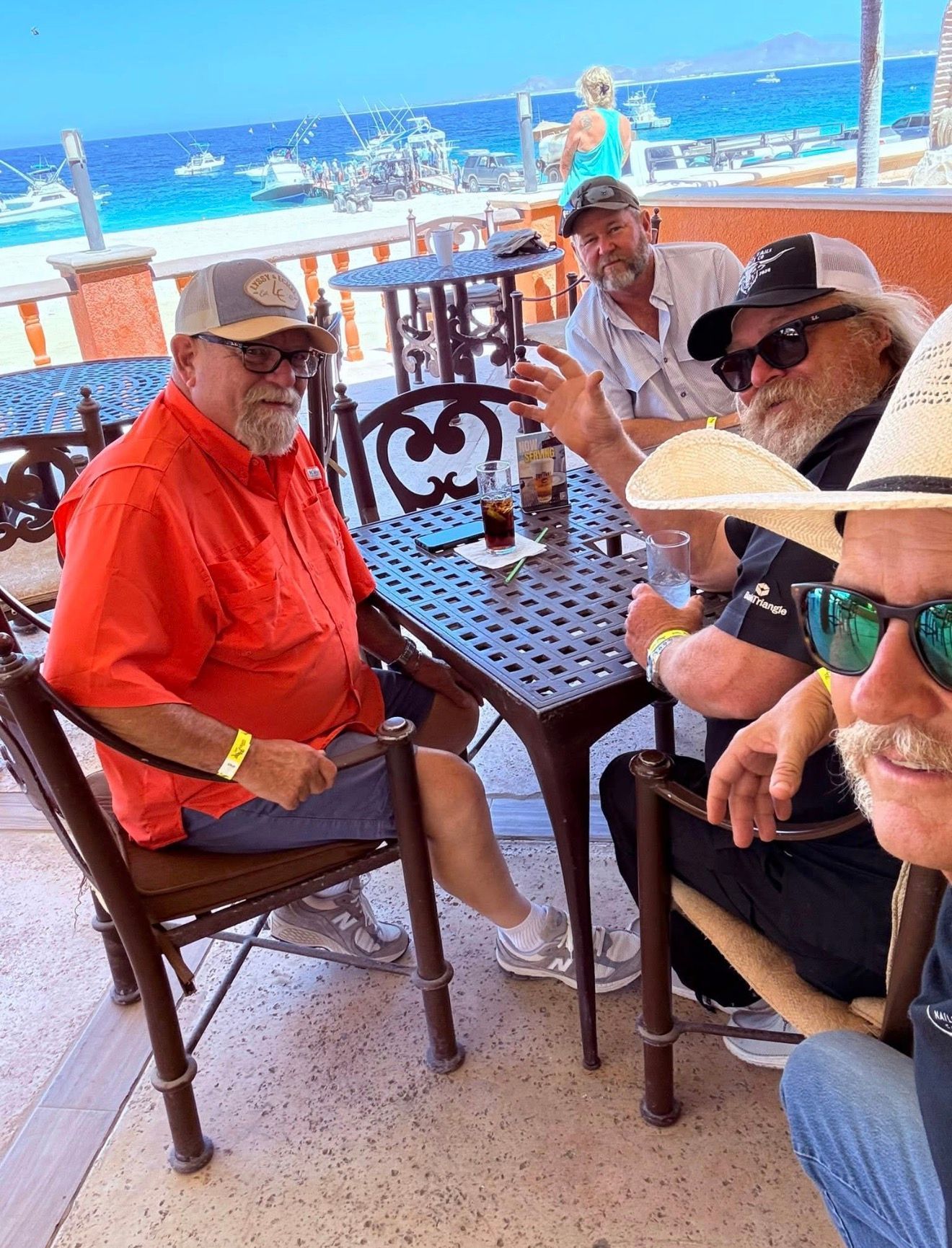 Four people sit at a seaside café table, chatting and smiling under umbrellas by the ocean.