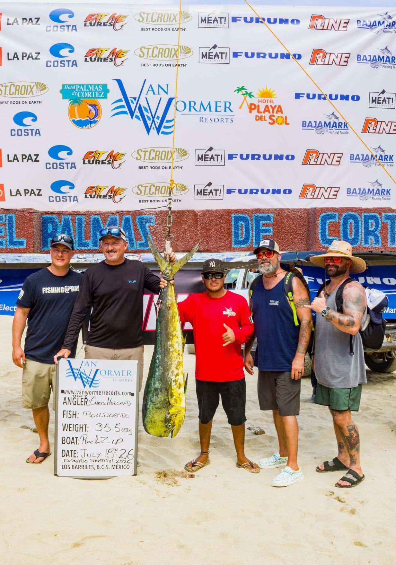 Five people on a beach hold a large fish trophy and sign in front of a sponsor backdrop.