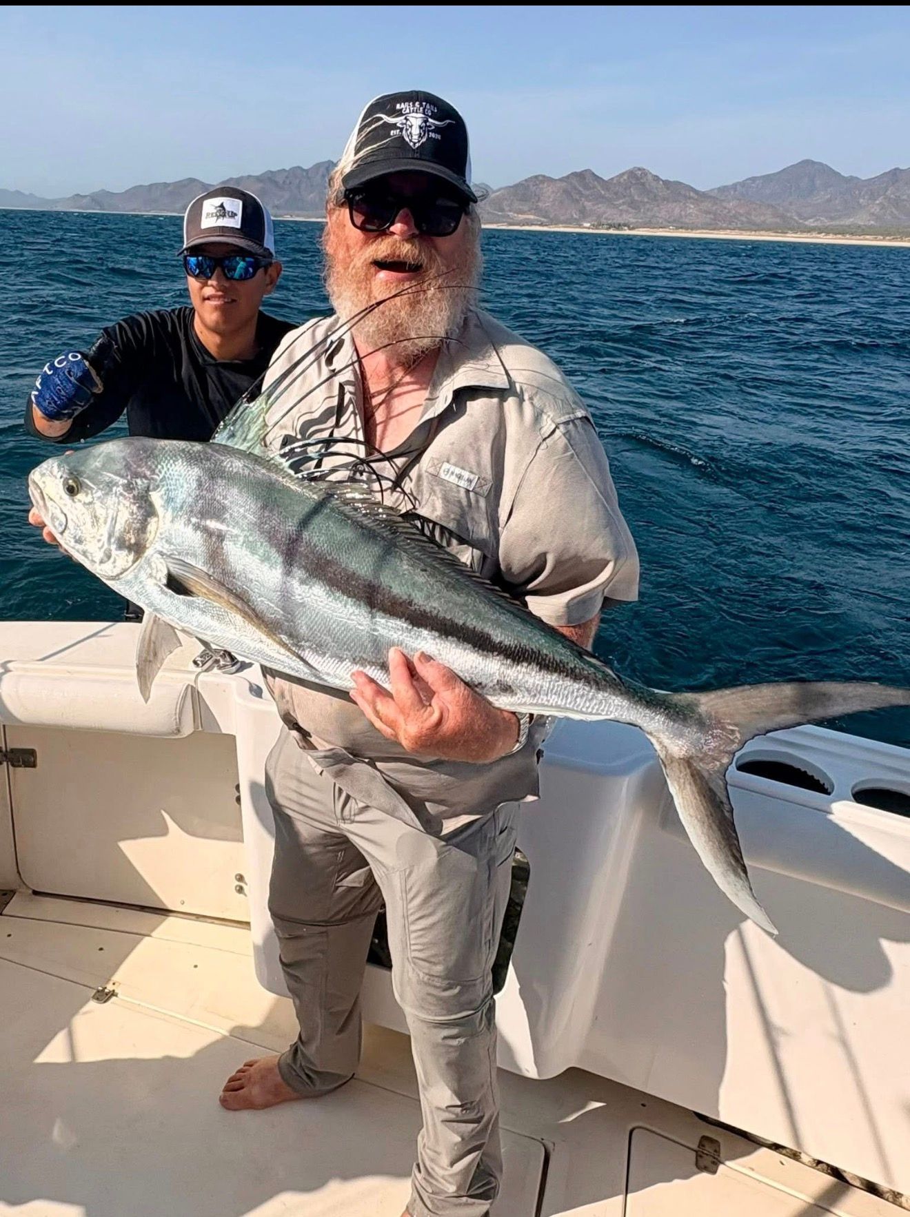 Two anglers on a boat holding a large silver fish on open water with mountains in the background