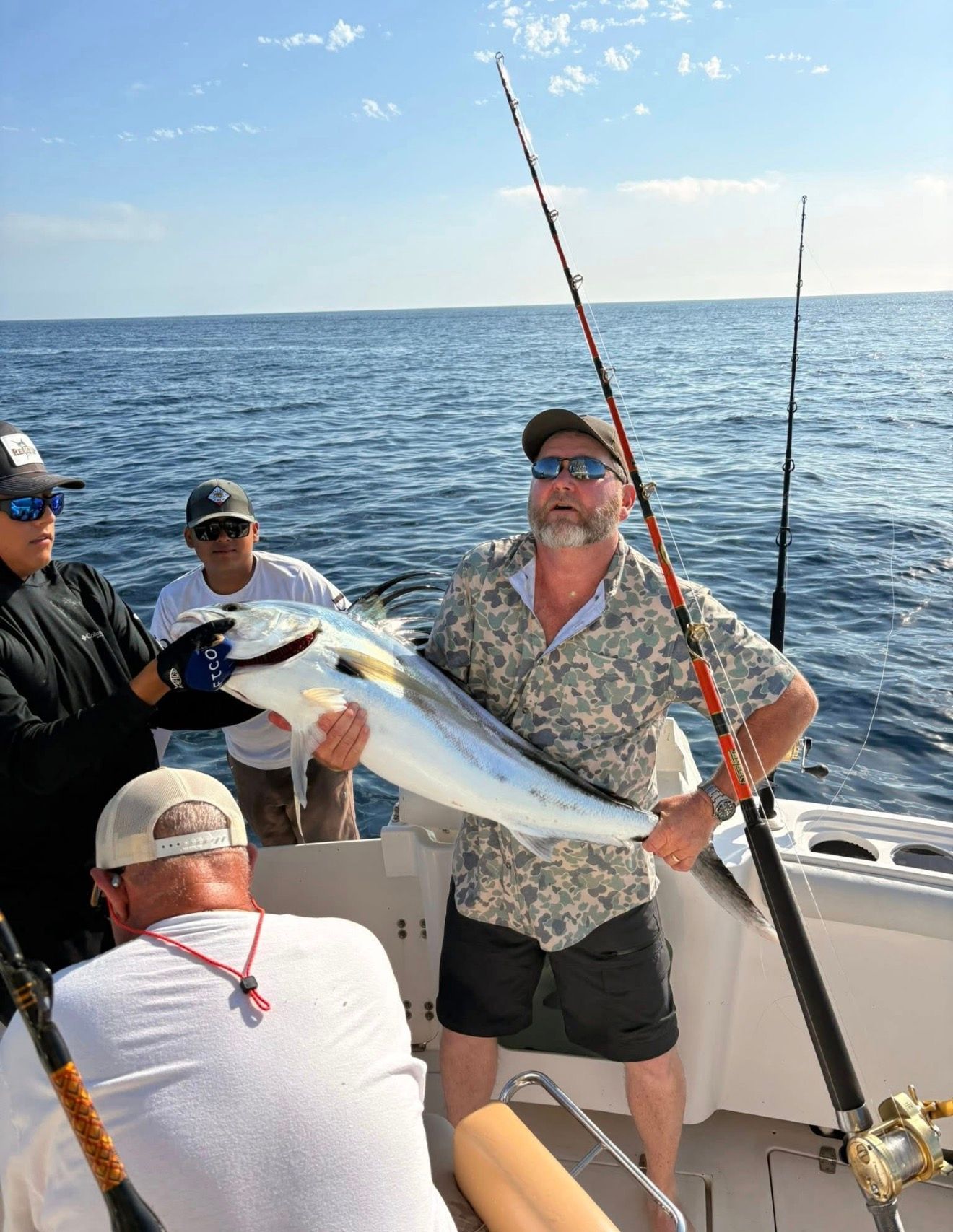 Anglers on a boat at sea holding a large fish, with fishing rods and calm blue water behind them.