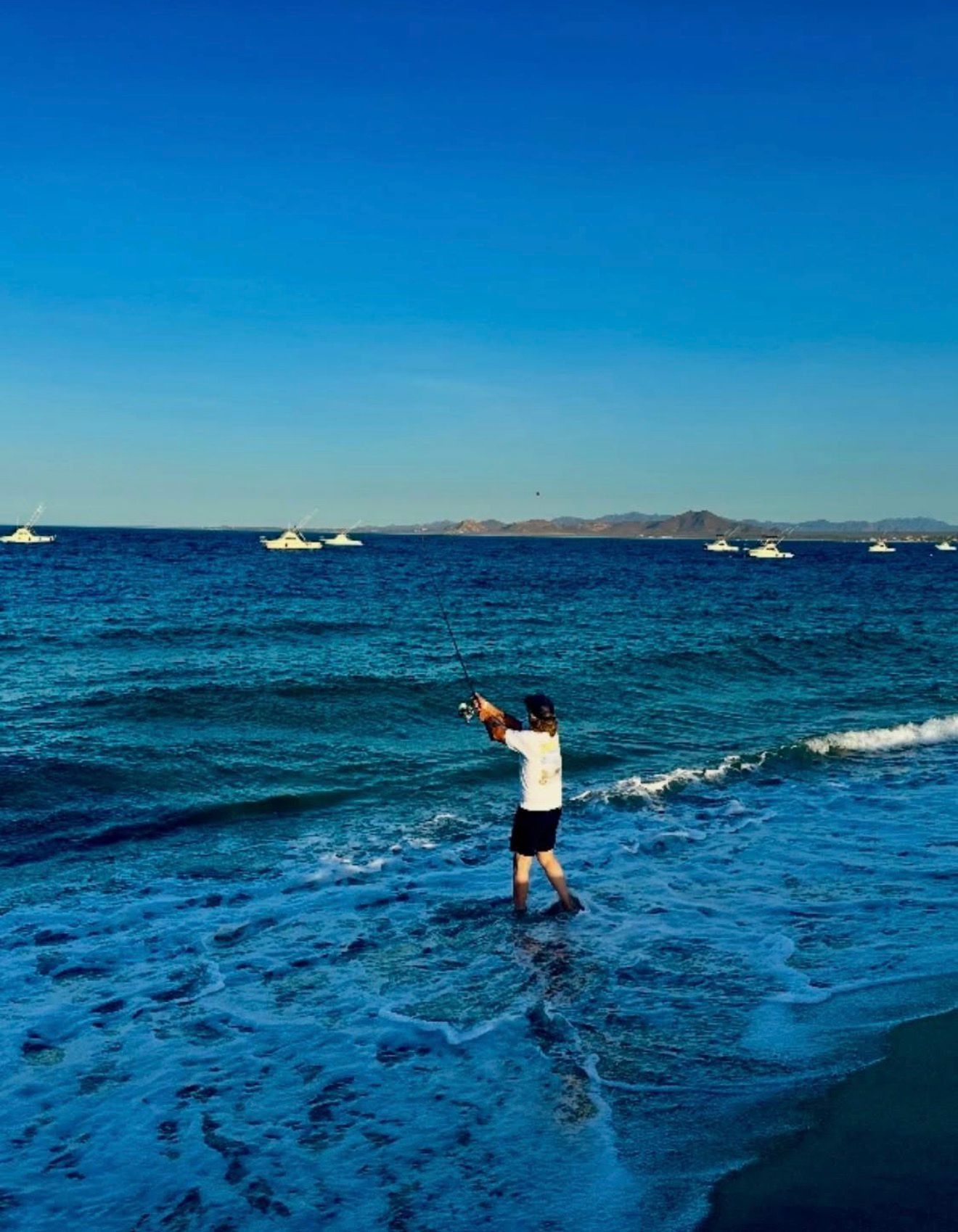 Person standing in shallow ocean water taking a photo, with blue sea and distant boats under a clear sky.