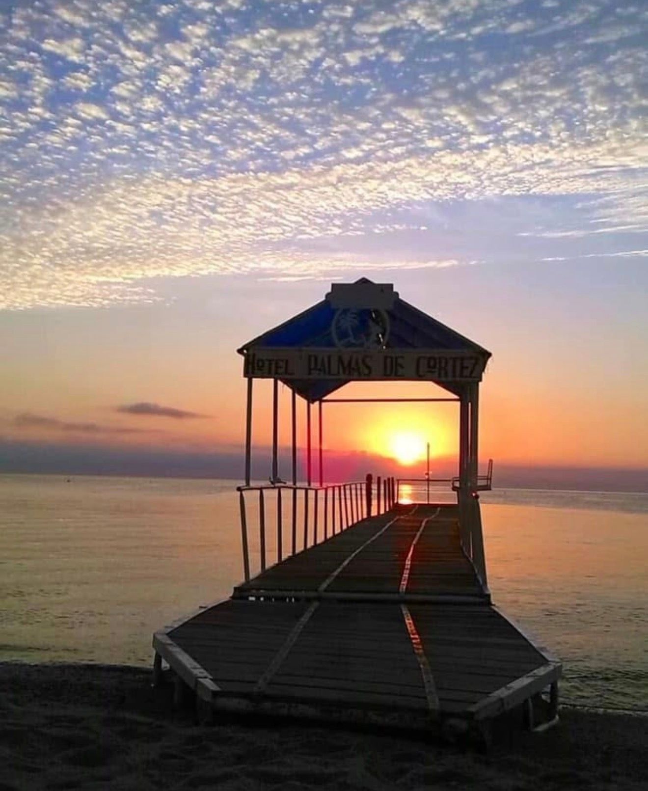 Sunset over a pier leading to a small gazebo, with calm water and a sky of scattered clouds.