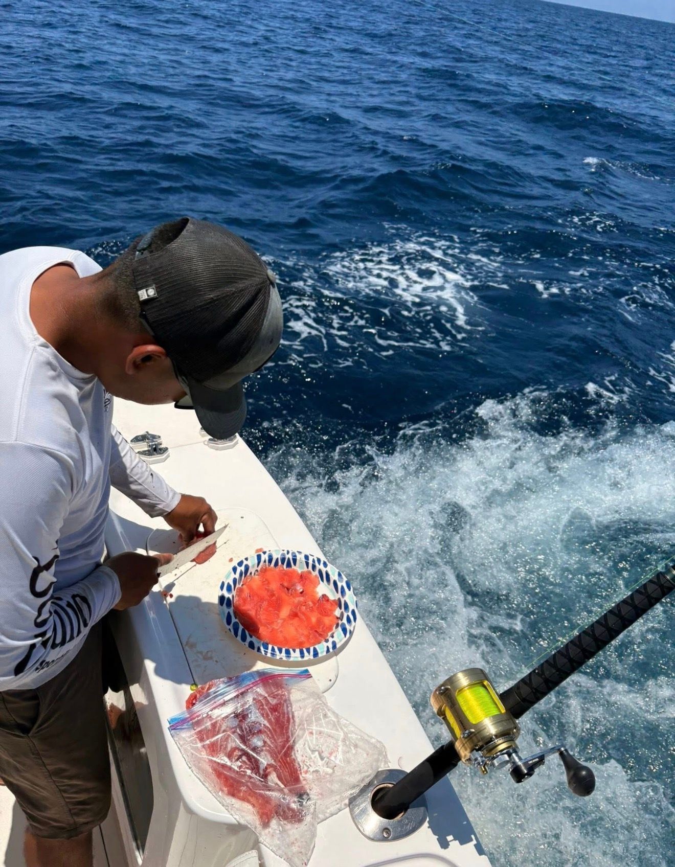 Person on a boat prepares food beside a blue ocean, with a plate of chopped tomatoes and a fishing rod nearby