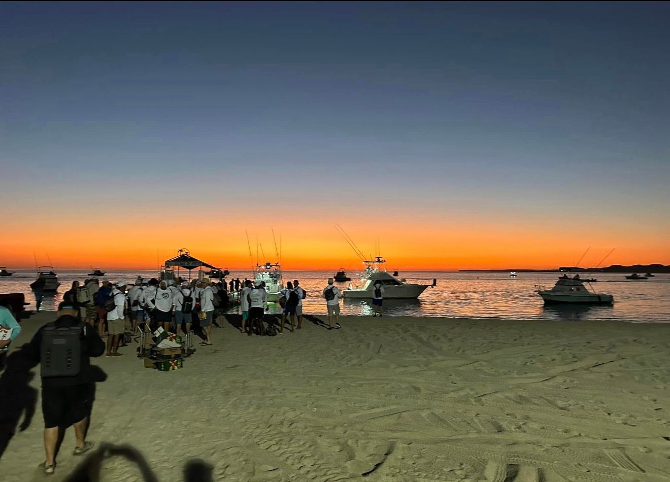 People walking on a beach at sunset with boats on the water and an orange-blue sky.