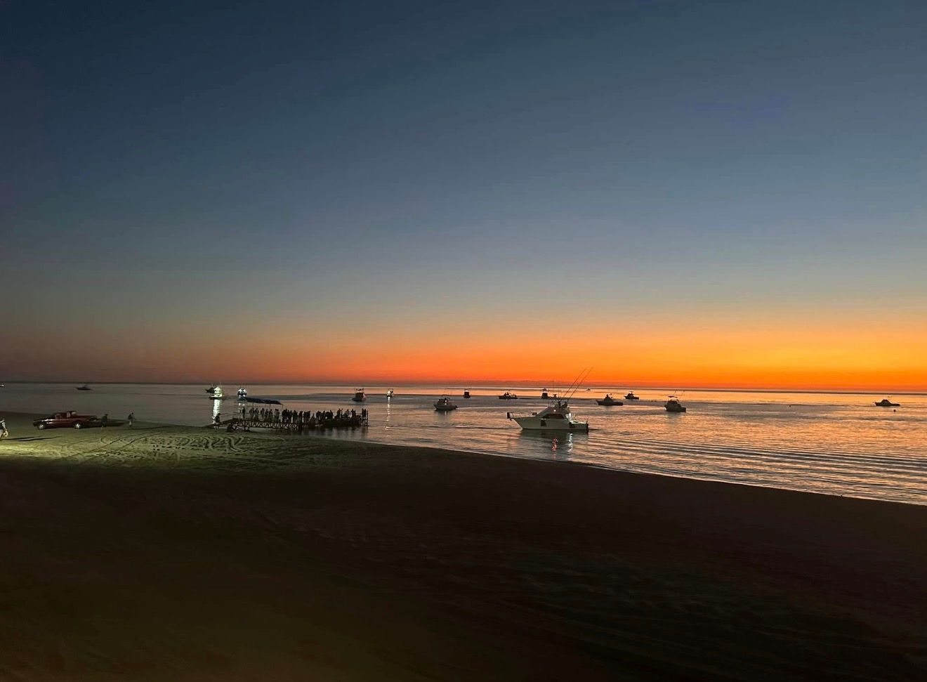 Sunset over a beach with boats on calm water and dark shoreline silhouettes