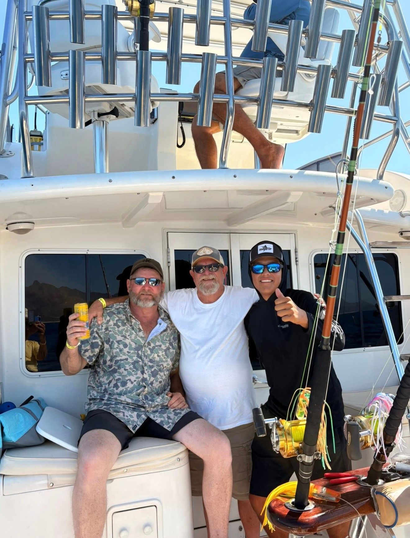 Three people smiling on a boat, holding drinks in bright sunlight.