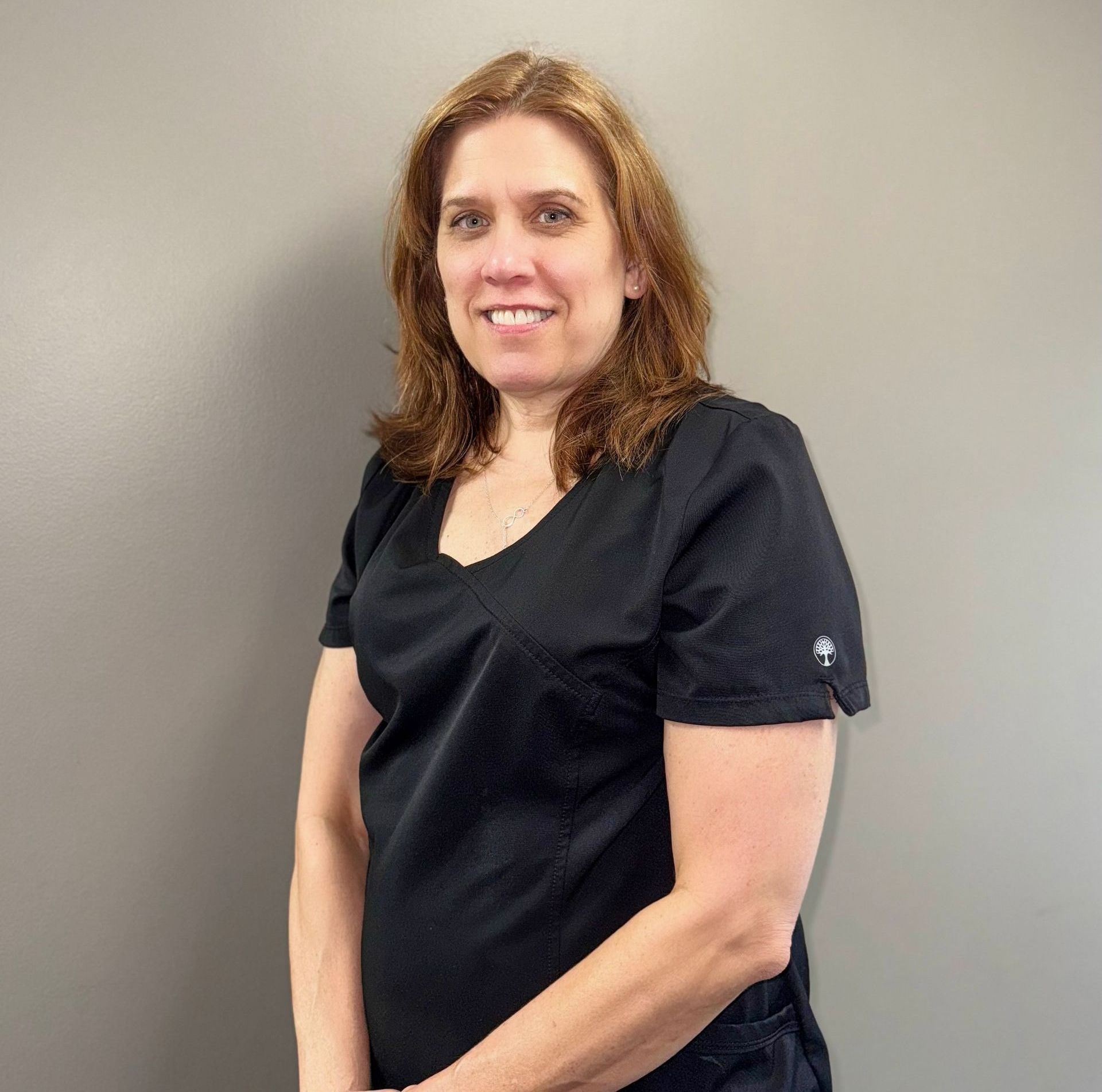 Woman in black scrubs smiles, standing against a gray wall.