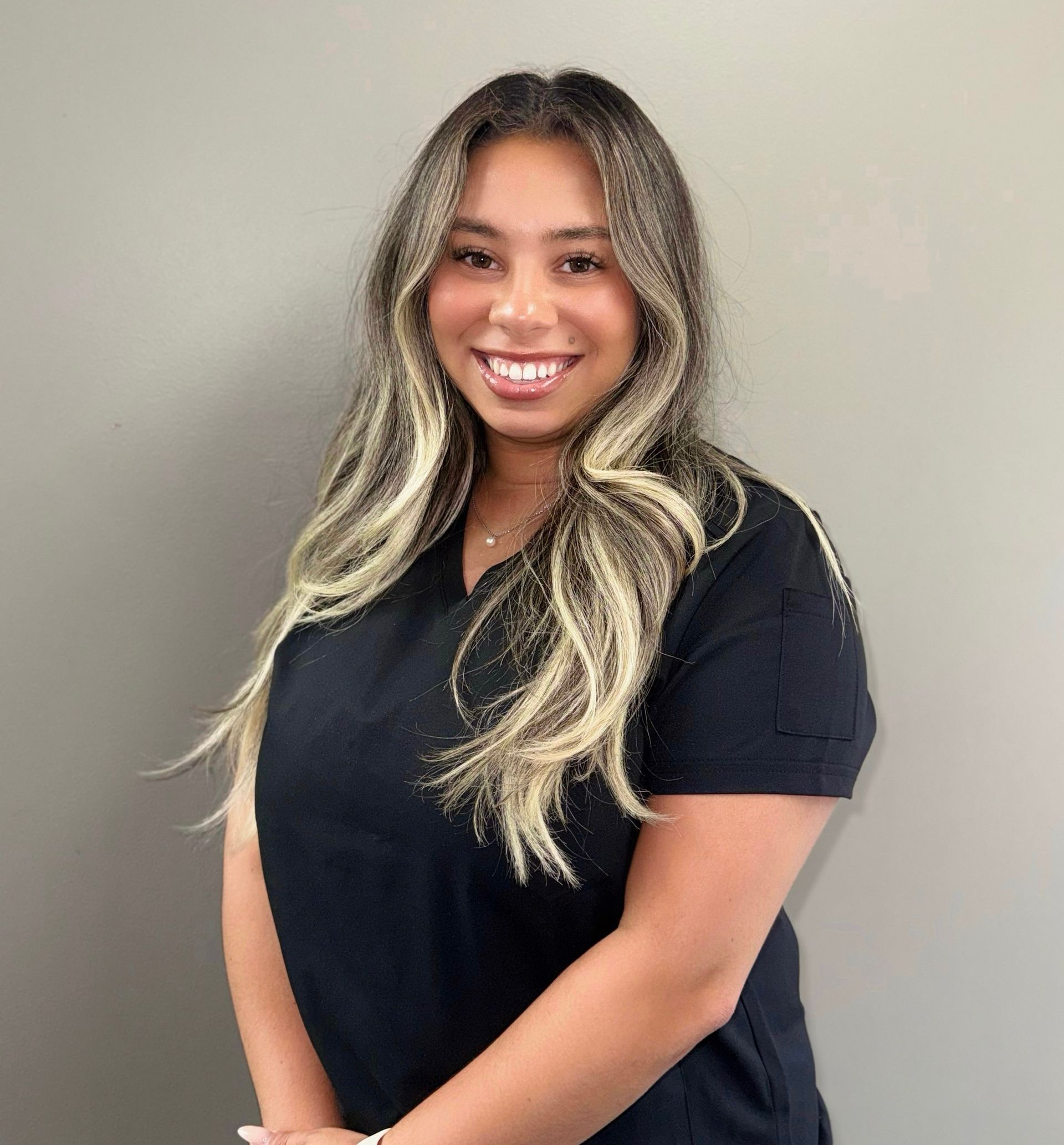 Woman with long, wavy blonde-highlighted hair smiles. She wears a black top in front of a gray wall.