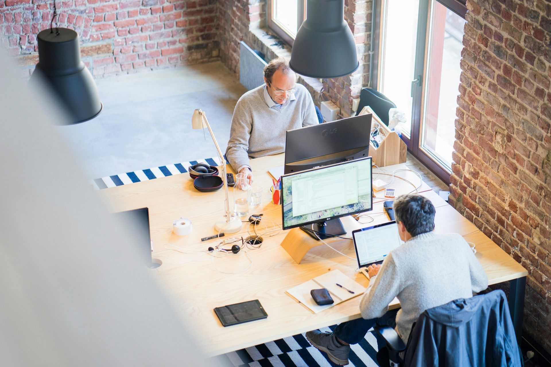 Two people working at a shared desk in a bright office with laptops and hanging lamps
