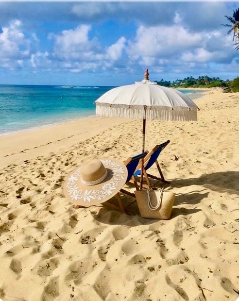 A wooden beach chair with a white fabric seat sits on the sand, featuring a red hat and sunglasses resting on the back. Beach chairs and straw hats under a white umbrella on a sunny tropical beach by the turquoise sea.