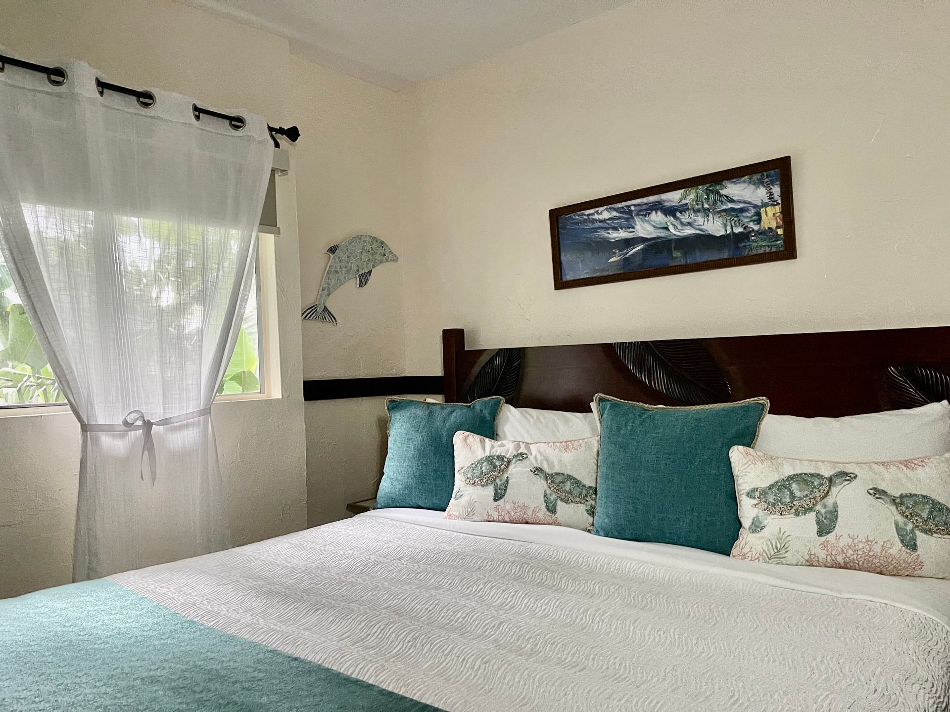 A coastal-themed bedroom with a bed featuring teal pillows, a dolphin wall decal, and a rectangular landscape painting.