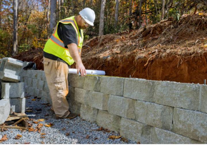 A man wearing a safety vest and hard hat builds a retaining wall, with trees in the background.