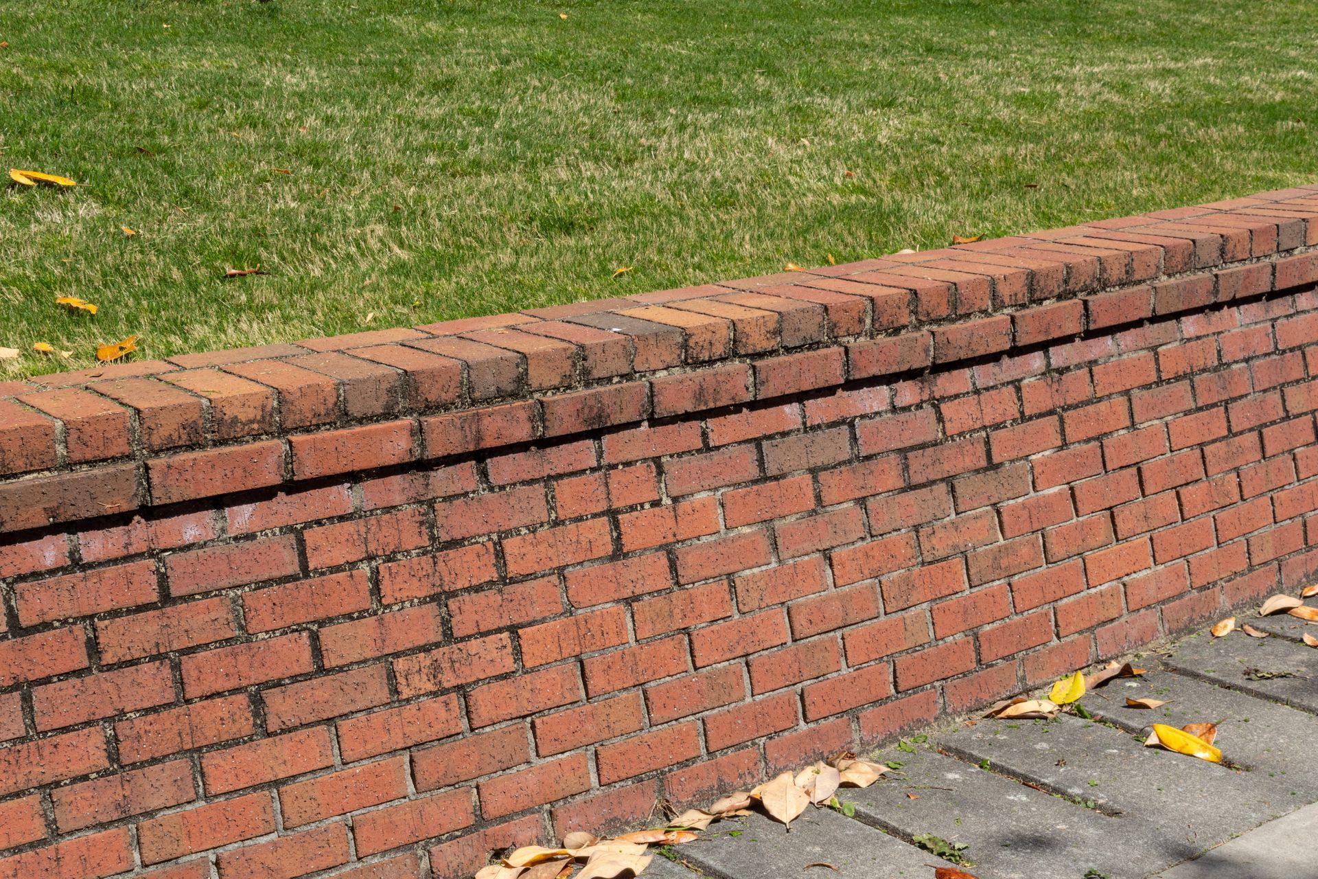 Brick retaining wall beside grass and a paved path, with yellow leaves on the ground.
