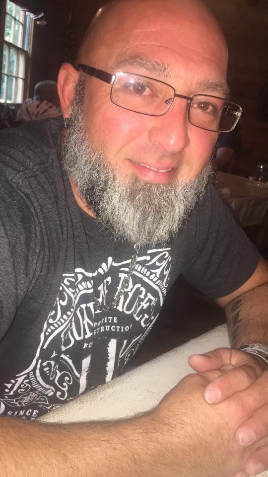 A person with a grey beard and glasses wearing a black t-shirt, sitting at a table with hands clasped.