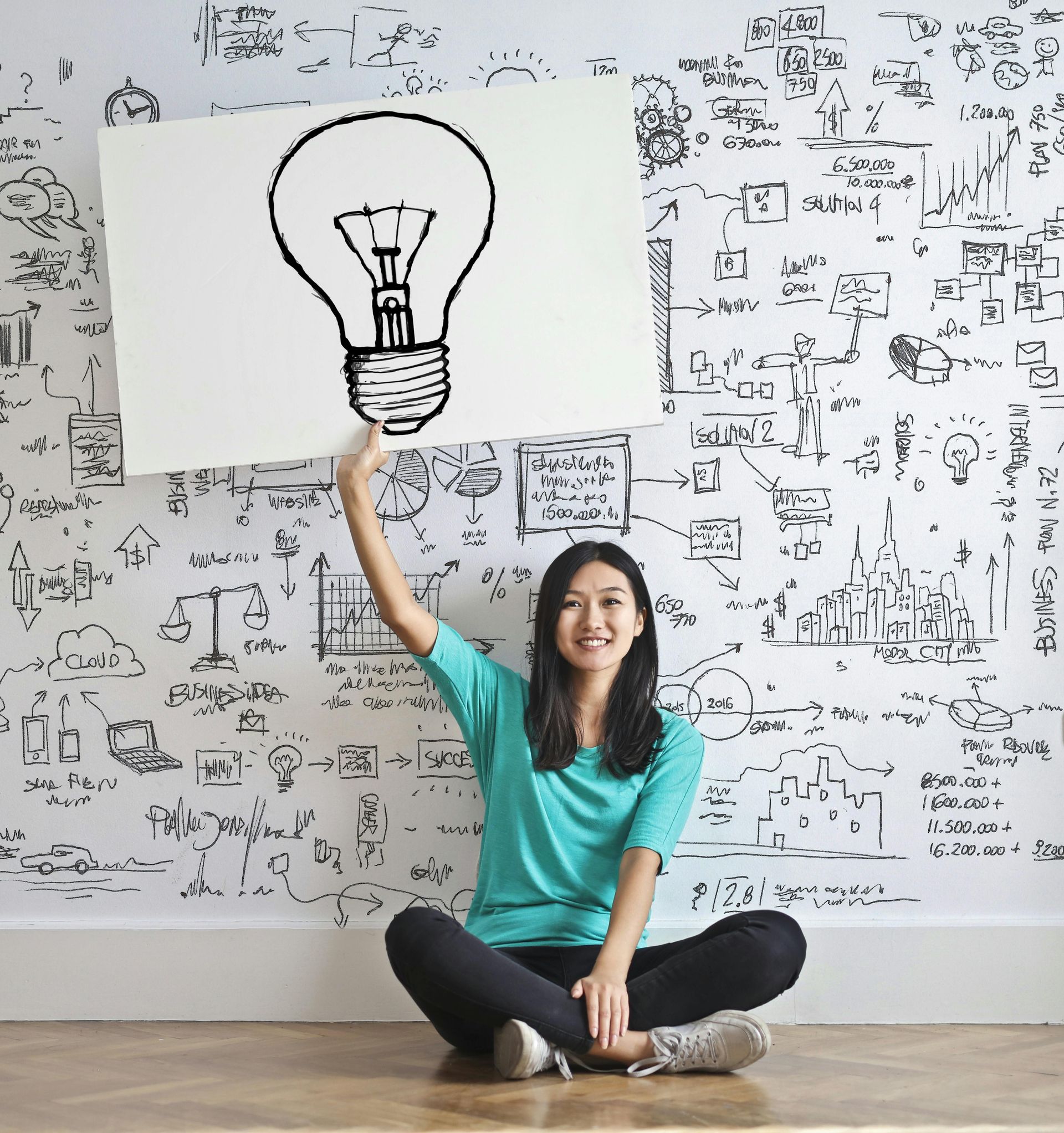 A woman is sitting on the floor holding up a sign with a light bulb drawn on it