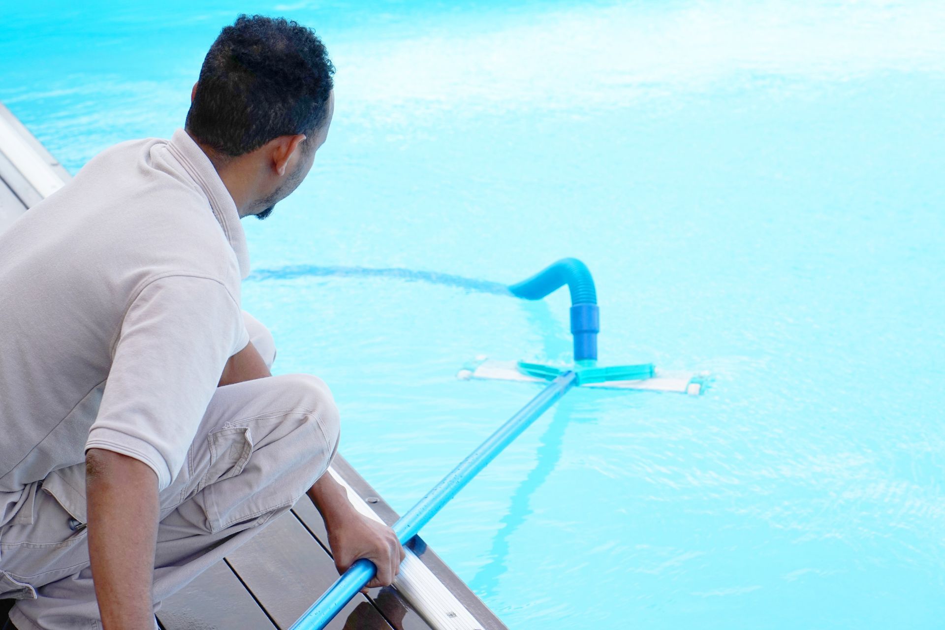 A person in a light-colored uniform uses a long pole and attachment to clean a bright blue swimming pool.