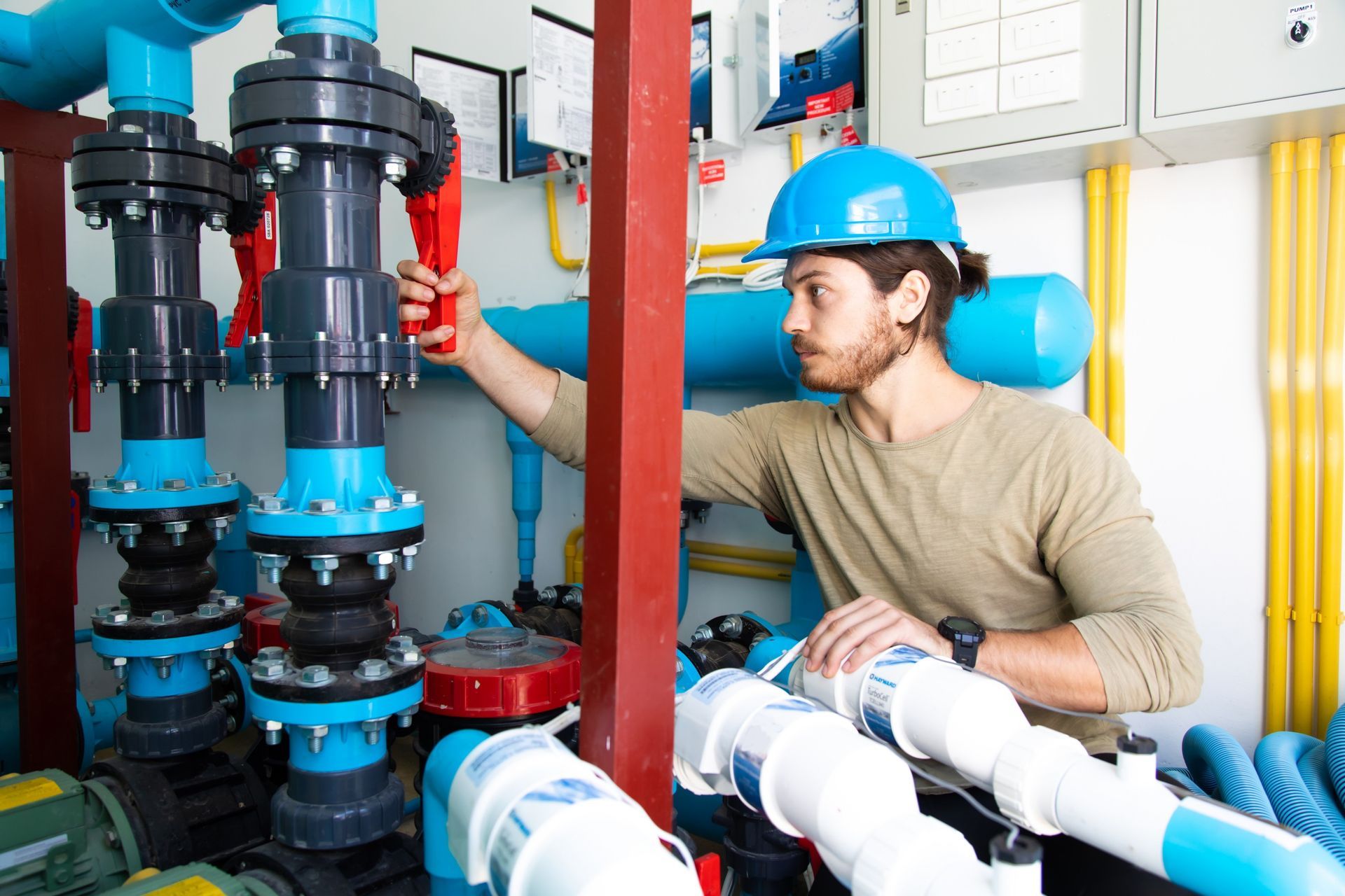 A person in a blue hard hat adjusting a red valve on a complex system of blue and white industrial pipes indoors.