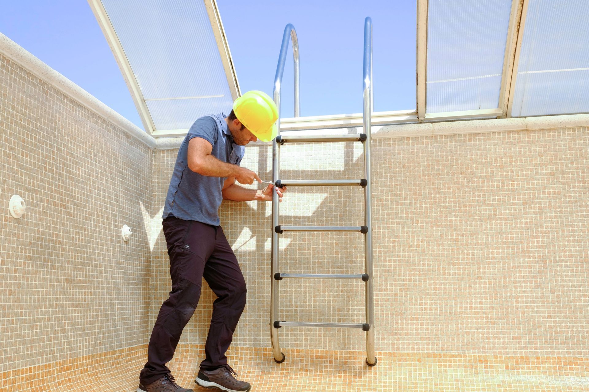 A person in a yellow hard hat and work clothes examines a stainless steel pool ladder inside a tiled pool.