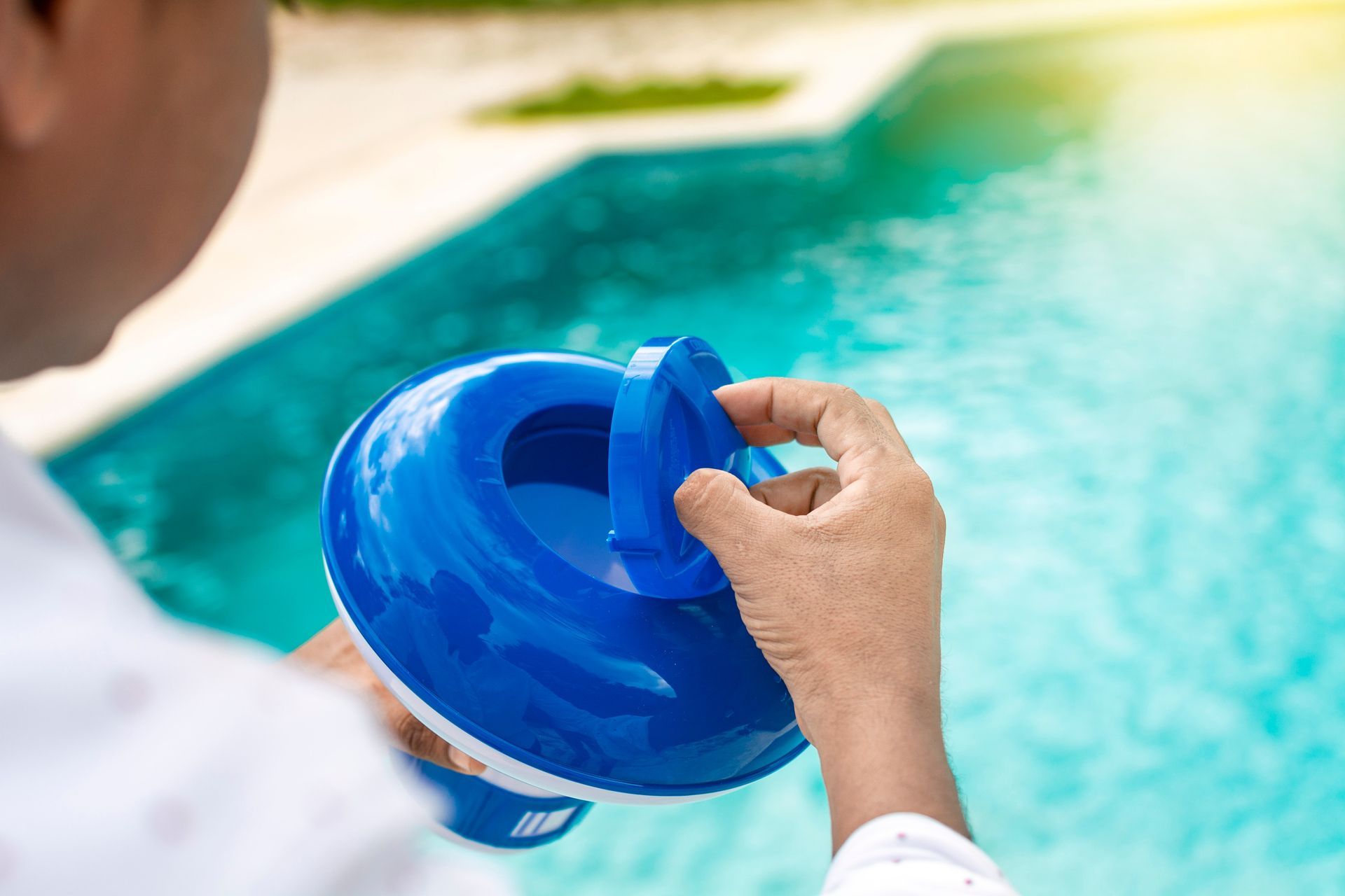 A person opens the blue lid of a floating pool chemical dispenser beside a swimming pool.