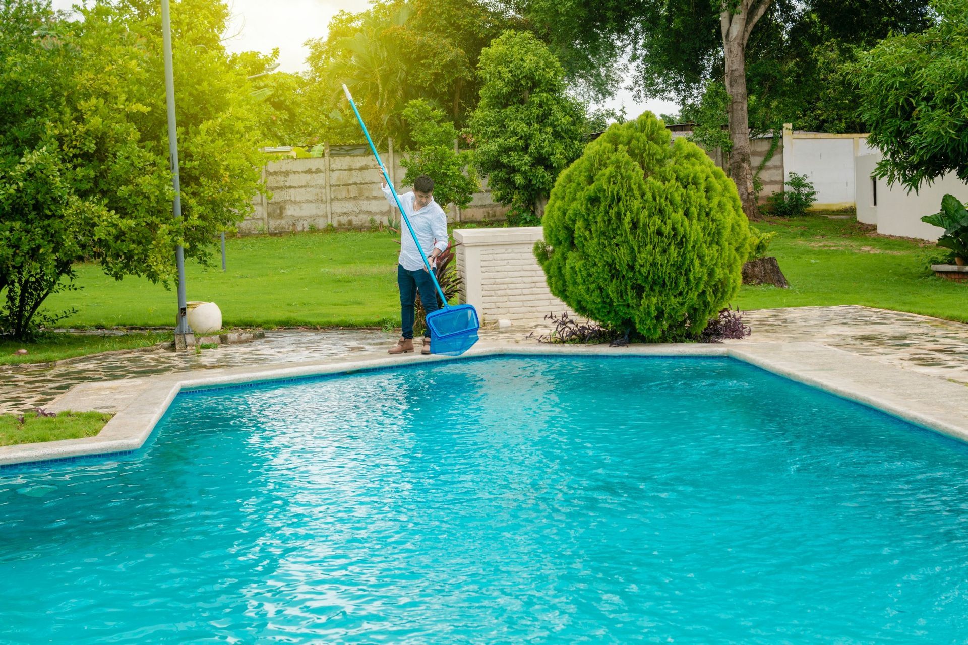 A person cleaning a bright blue backyard swimming pool with a long-handled net on a sunny day.