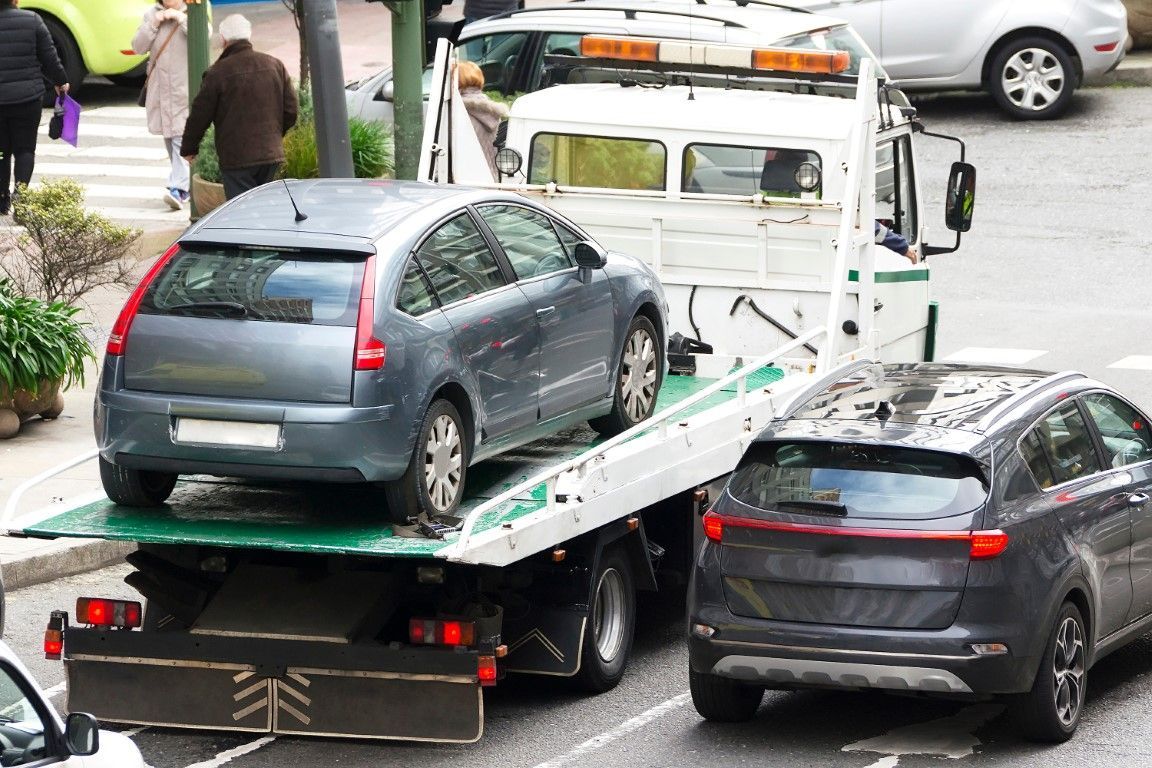 A gray car being towed by a tow truck on a street, with another car nearby and people walking.