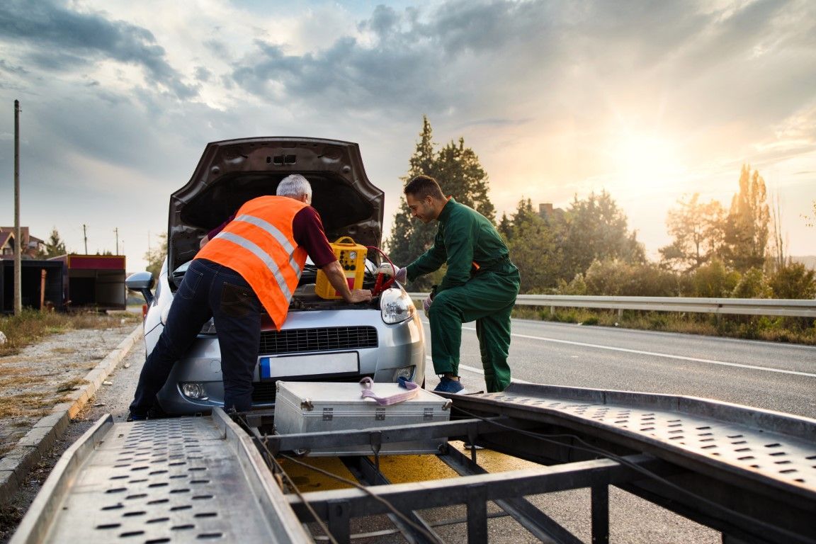 Two people loading a car onto a tow truck on the side of a road. One wears an orange vest.