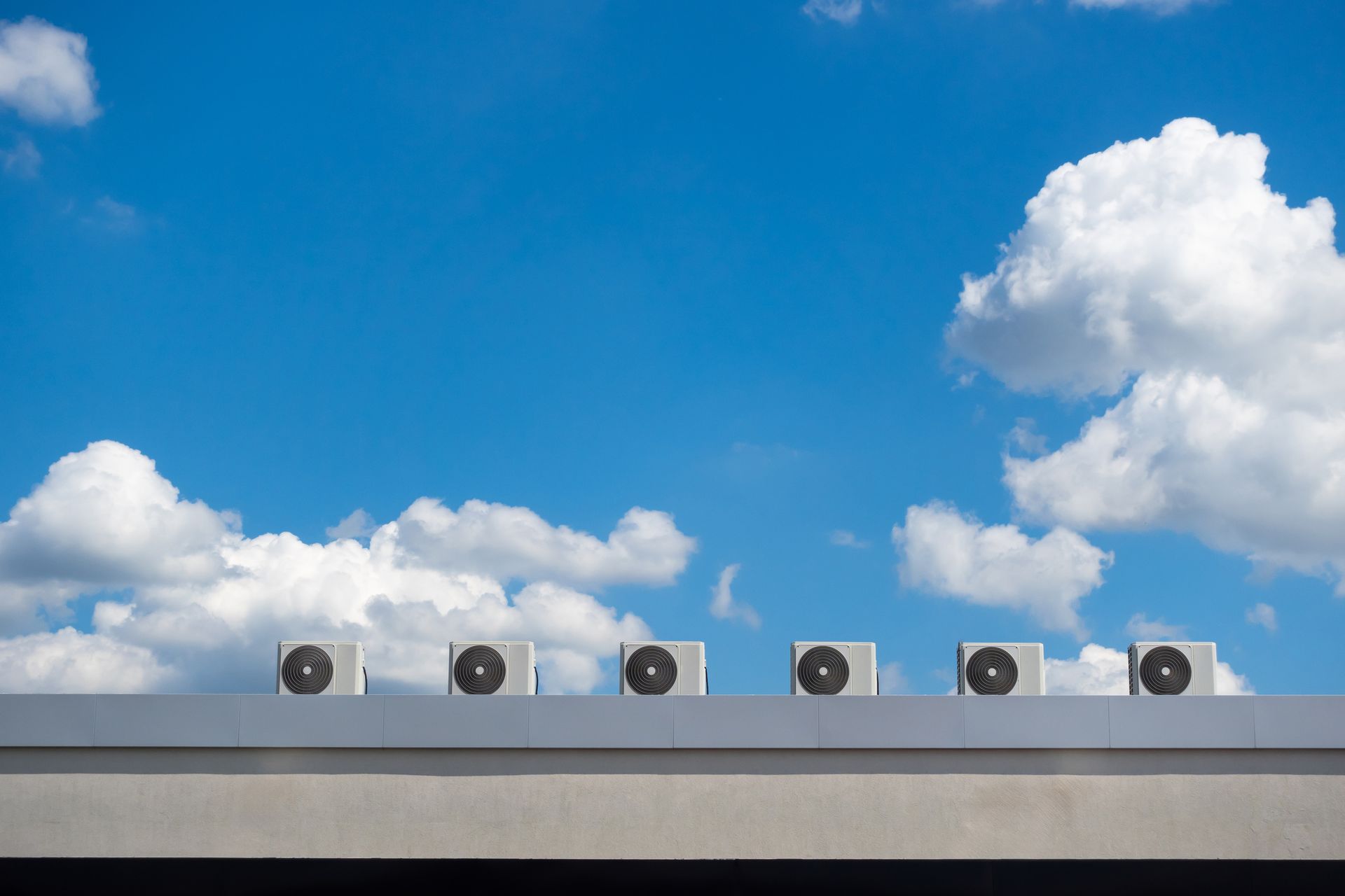 A row of six HVAC units sit atop a flat roof against a bright blue sky filled with white clouds.