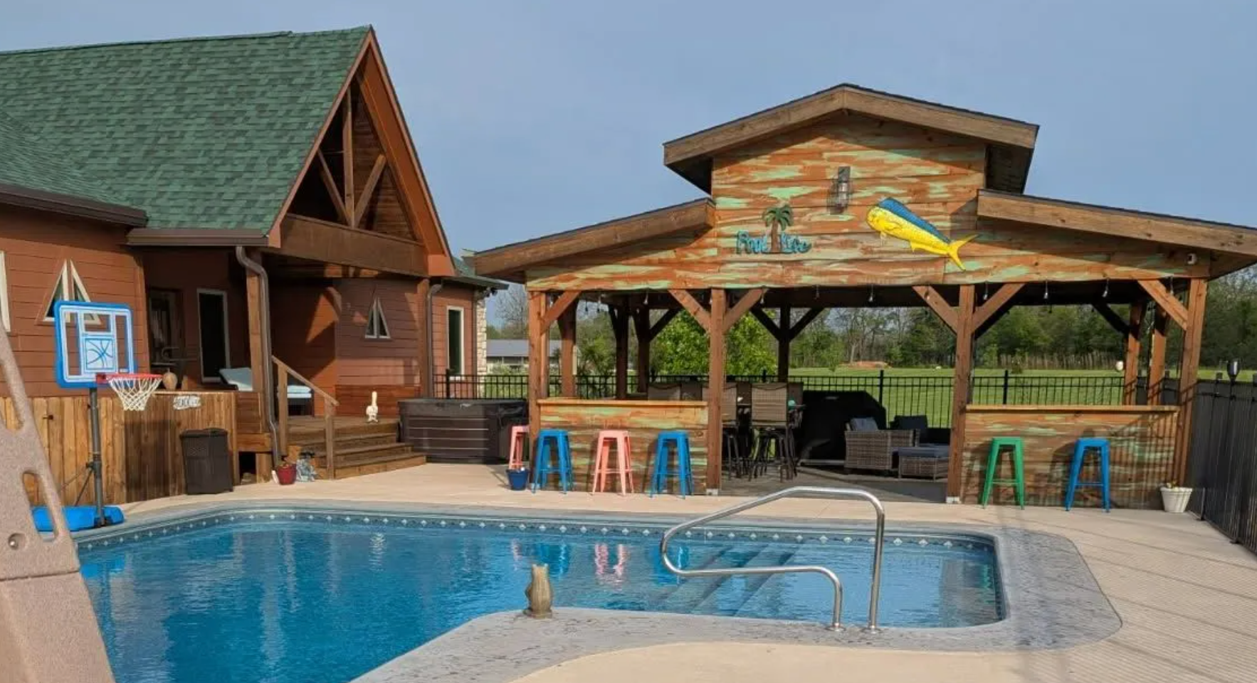 Wooden lodge and shaded patio beside a blue swimming pool with lounge chairs and a hot tub