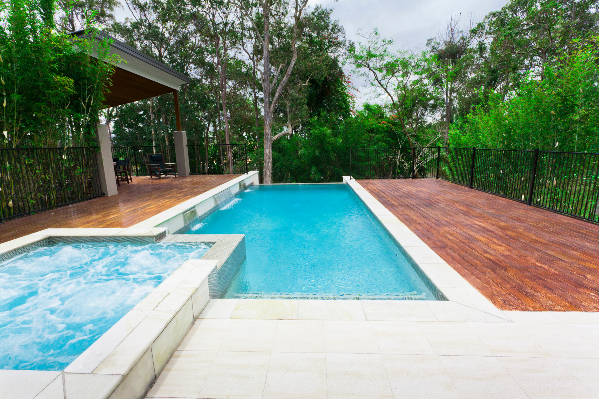 Modern backyard pool with hot tub, turquoise water, and wooden deck surrounded by lush greenery