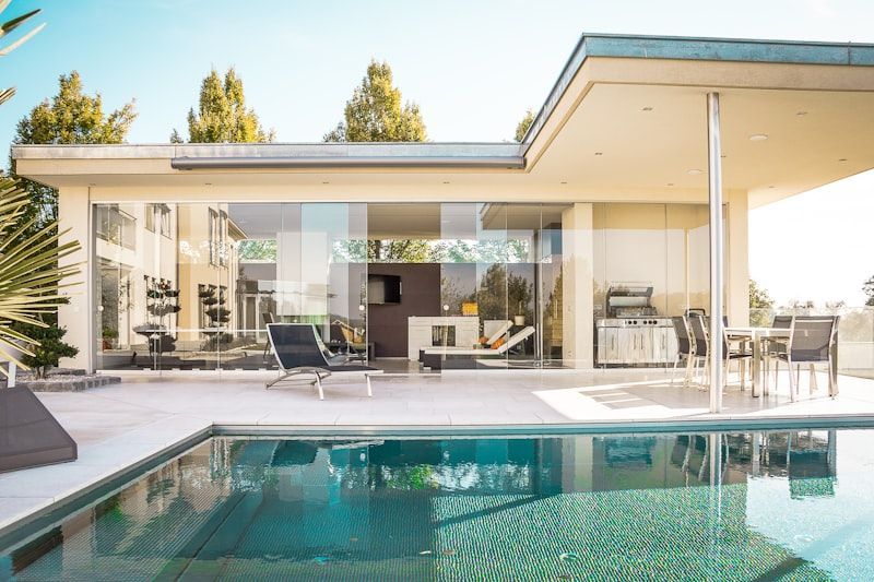 Modern home patio with glass walls and turquoise swimming pool in the foreground