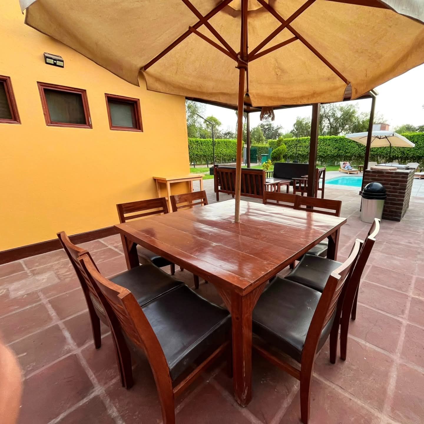 Outdoor dining table set with umbrella, near a pool, set against a yellow building.