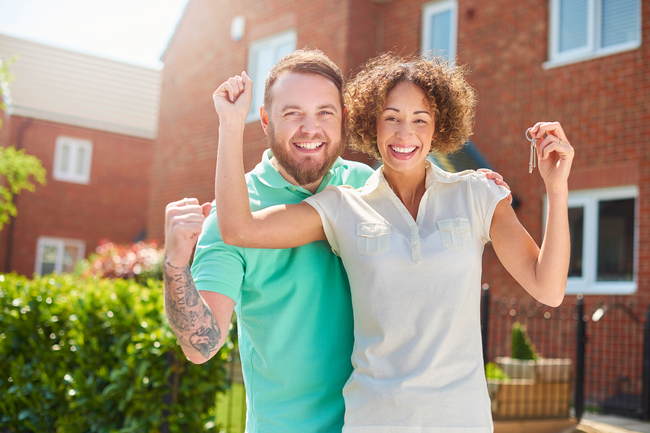 Smiling couple standing arm in arm outdoors in front of brick apartments in bright sunlight