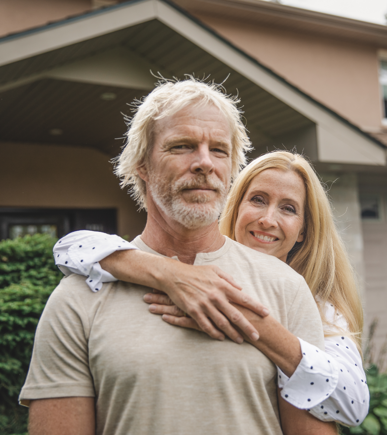 Two smiling adults standing outside a house, the woman hugging the man from behind.