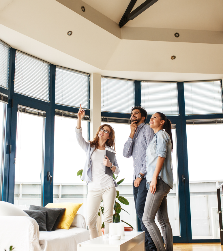 Three people chatting and smiling in a bright modern living room with large windows.