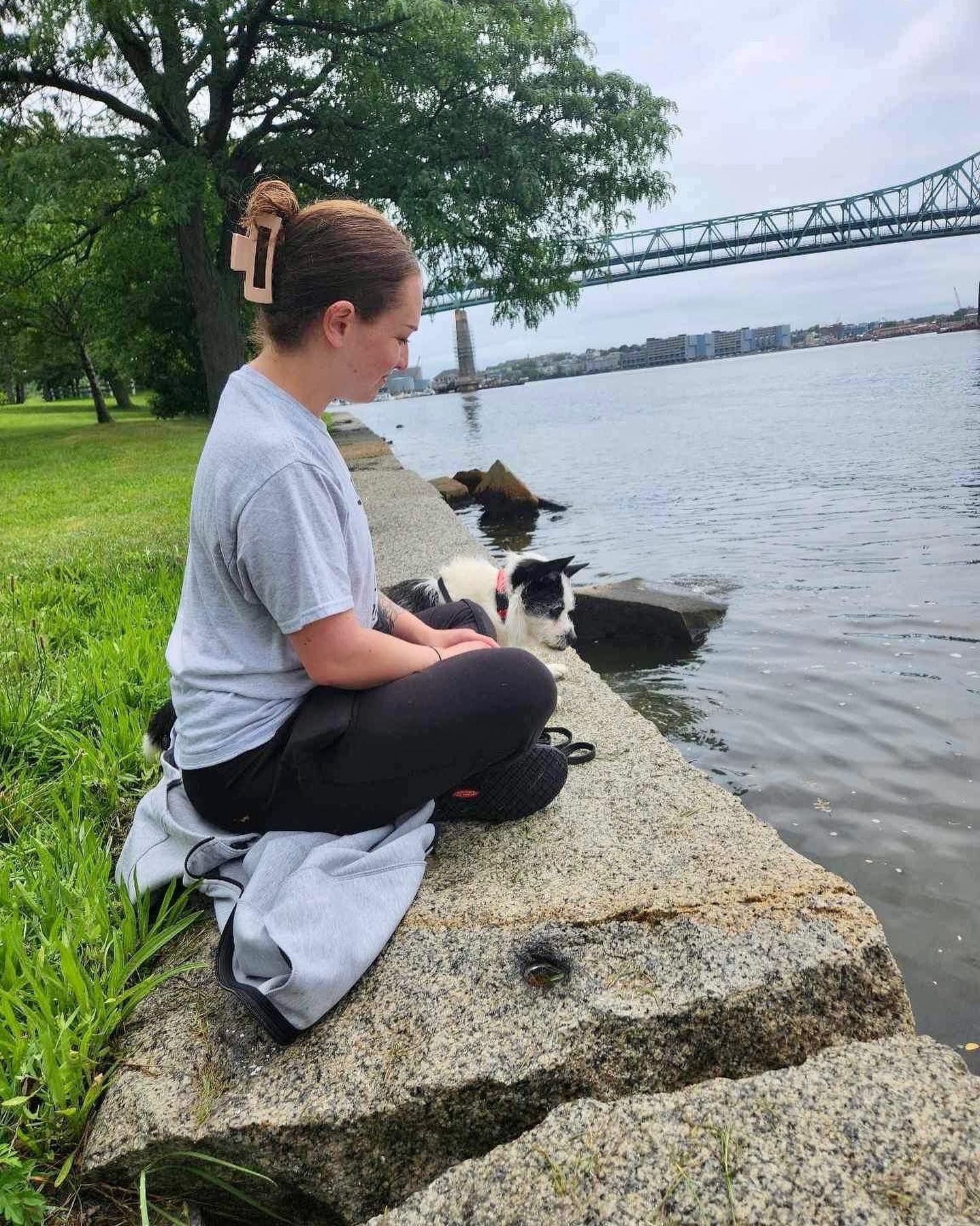A woman is sitting on a rock next to a body of water with a dog.