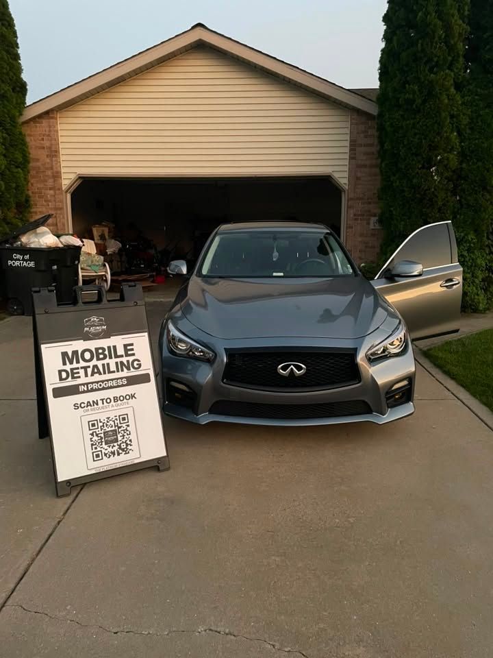 Gray Infiniti sedan in a driveway beside a garage, with a mobile detailing sign and open car door.