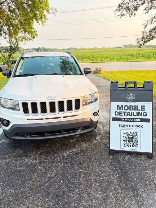 White SUV parked beside a mobile detailing sign on a driveway near a field at sunset