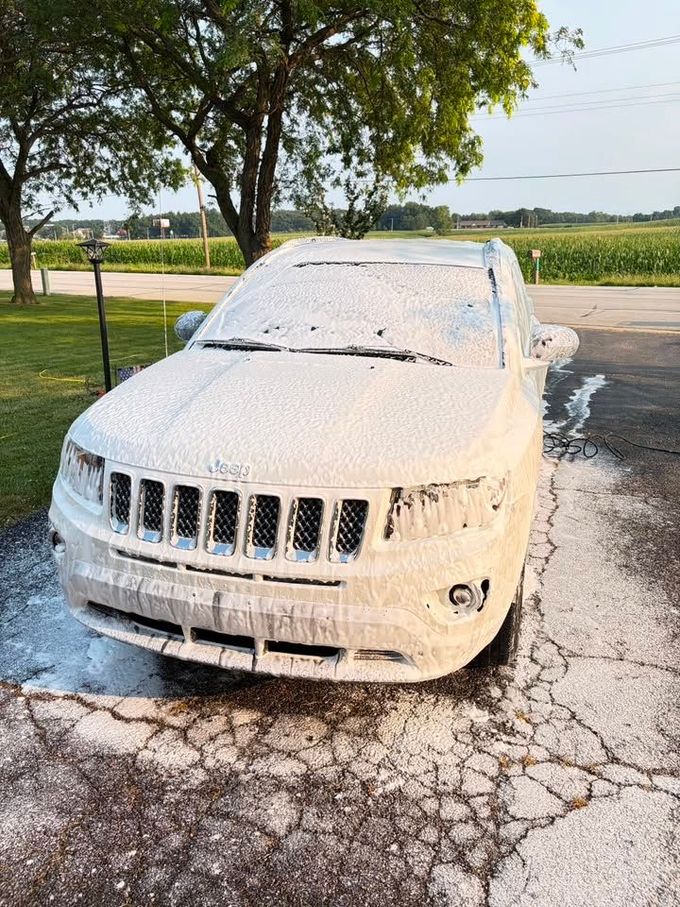 White SUV covered in foam, parked on cracked pavement by a grassy roadside with trees.