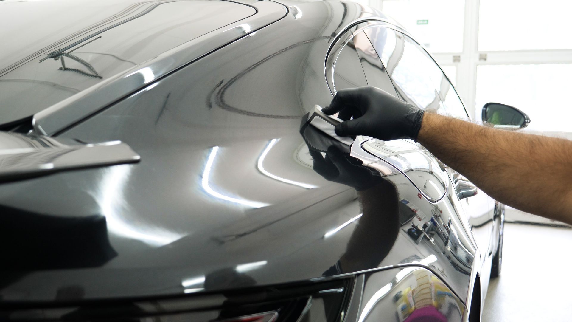 Gloved hand polishing a dark car hood in a bright garage