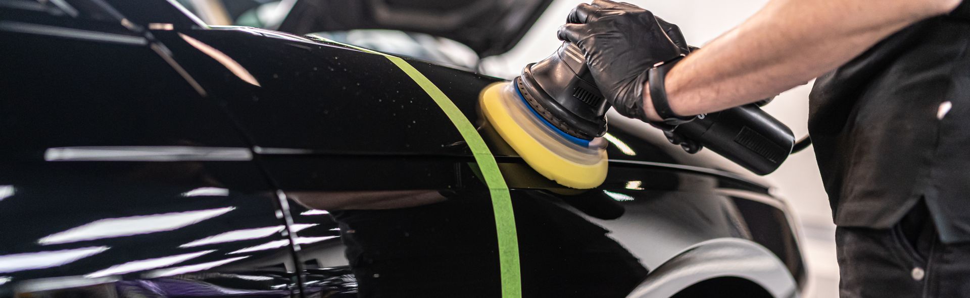 Person polishing a black car door with a green cloth and yellow buffer in a garage