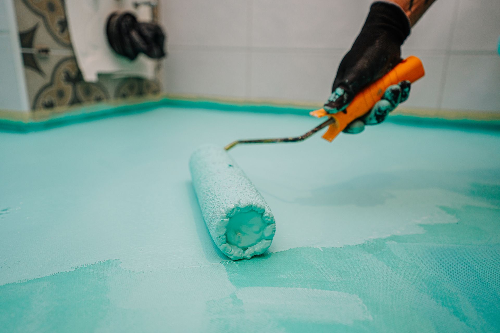 Hand using orange-handled paint roller to apply mint-green paint on a floor or surface