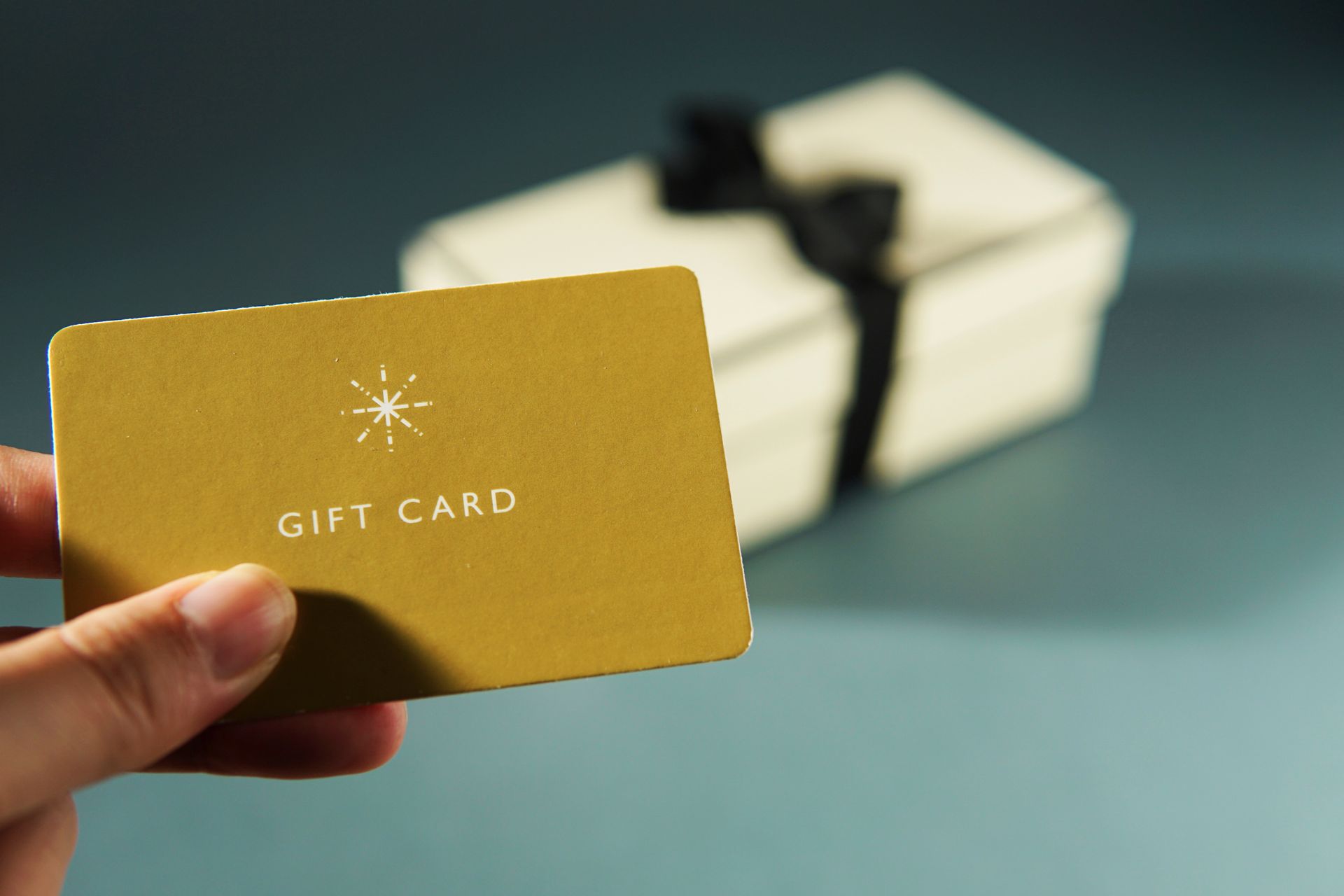 Hand holding a gold gift card, with a wrapped present blurred in the background.