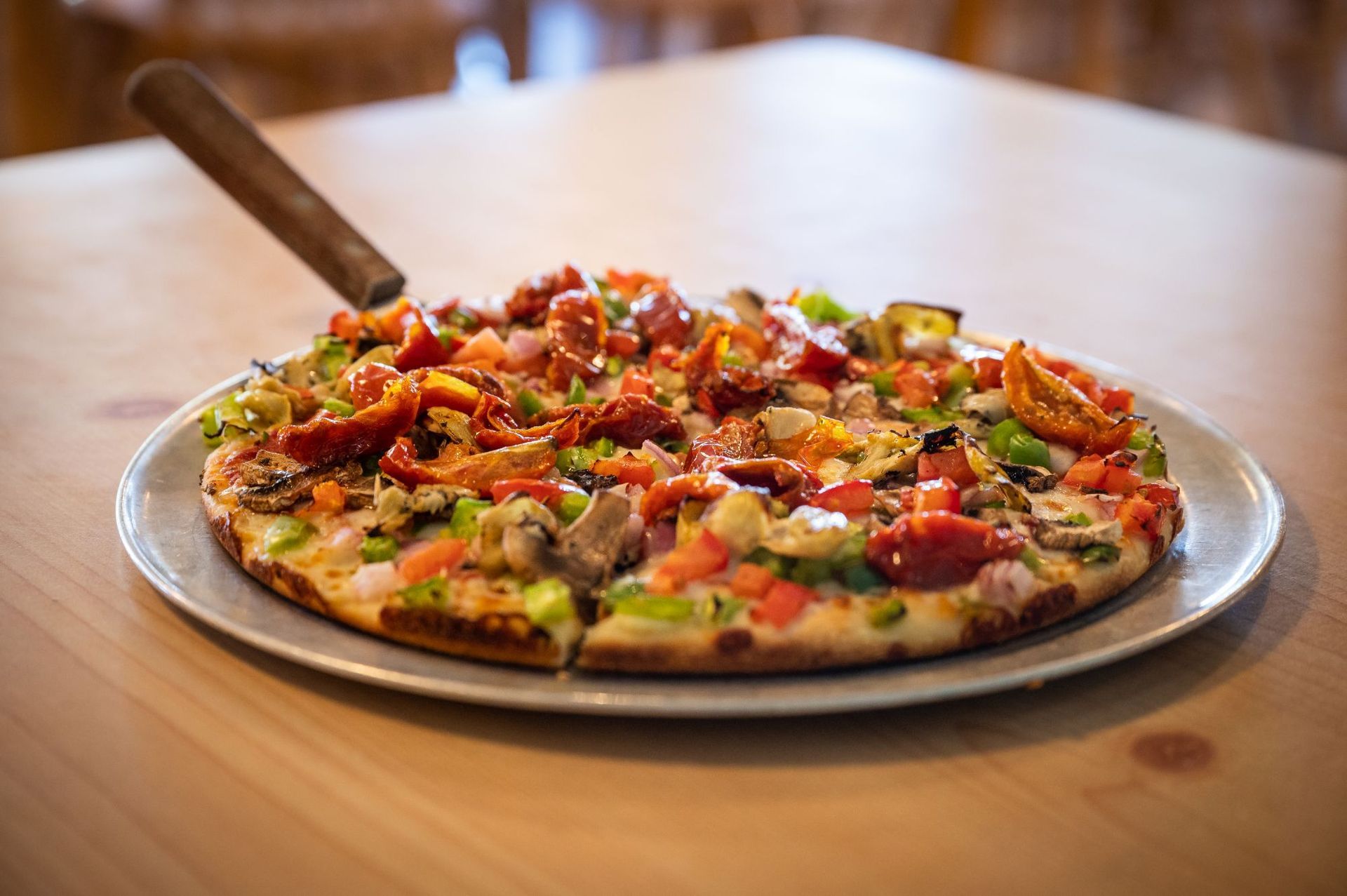 Pizza on a wooden table with a knife, topped with colorful vegetables and cheese