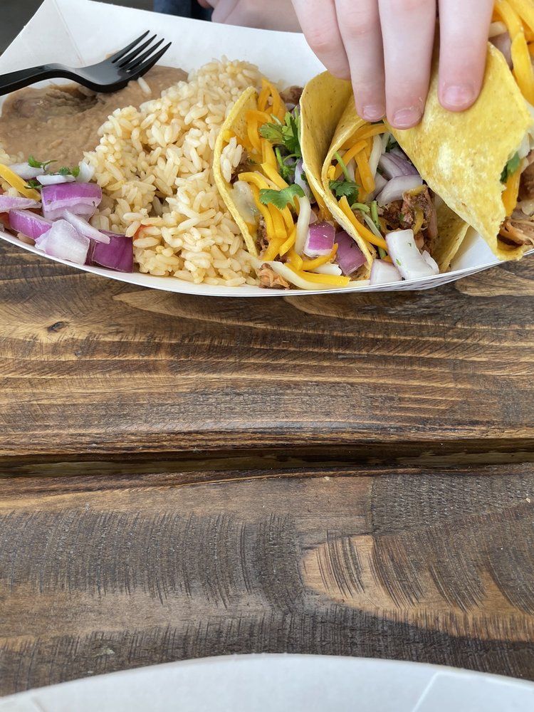 Hand lifting a taco over a plate of rice, tacos, and red onions on a wooden table