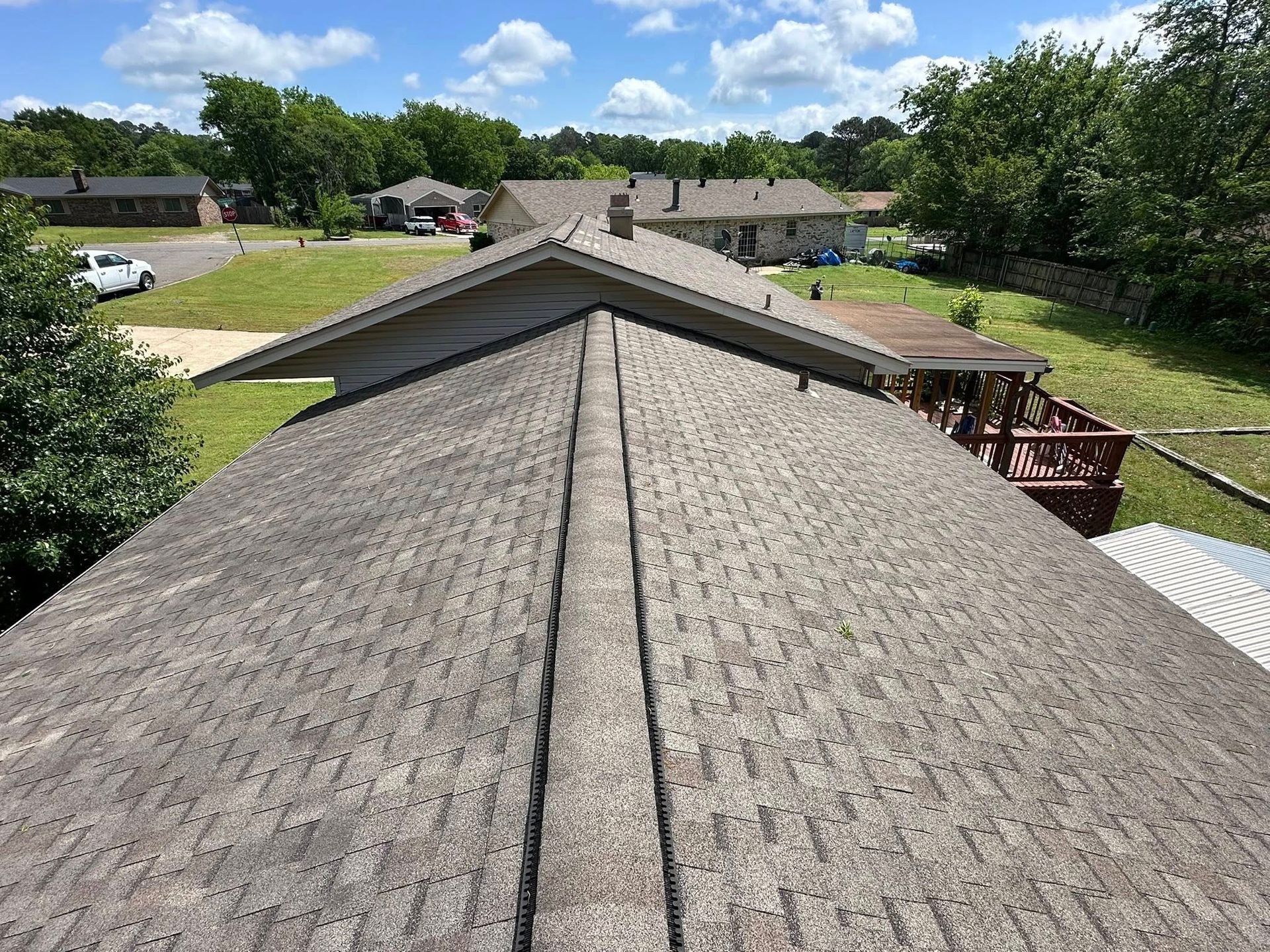 Rooftop view of gray weathered shingles with houses and green trees in the background under a blue sky.
