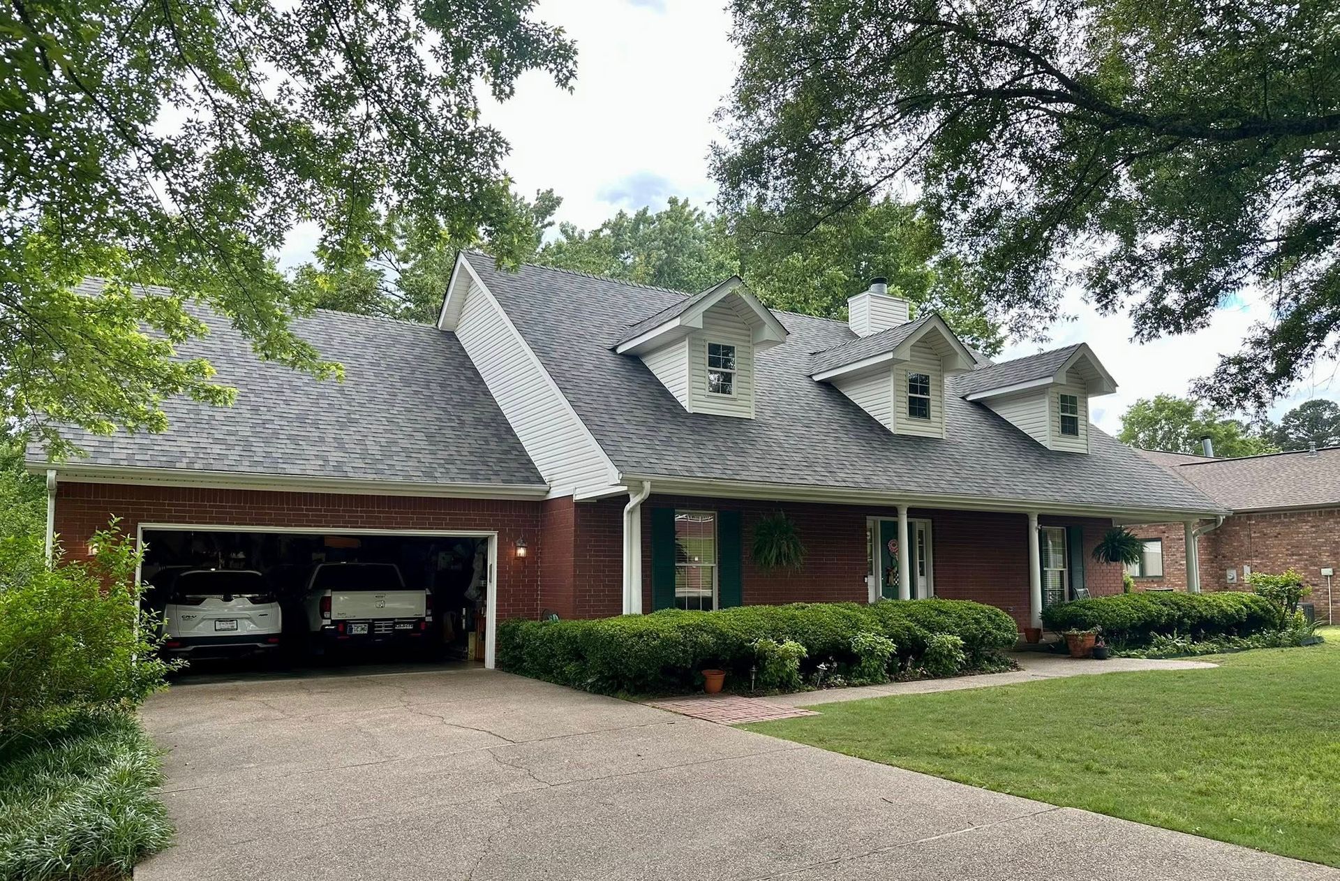 Red brick house with attached garage and dormers; cars parked inside.