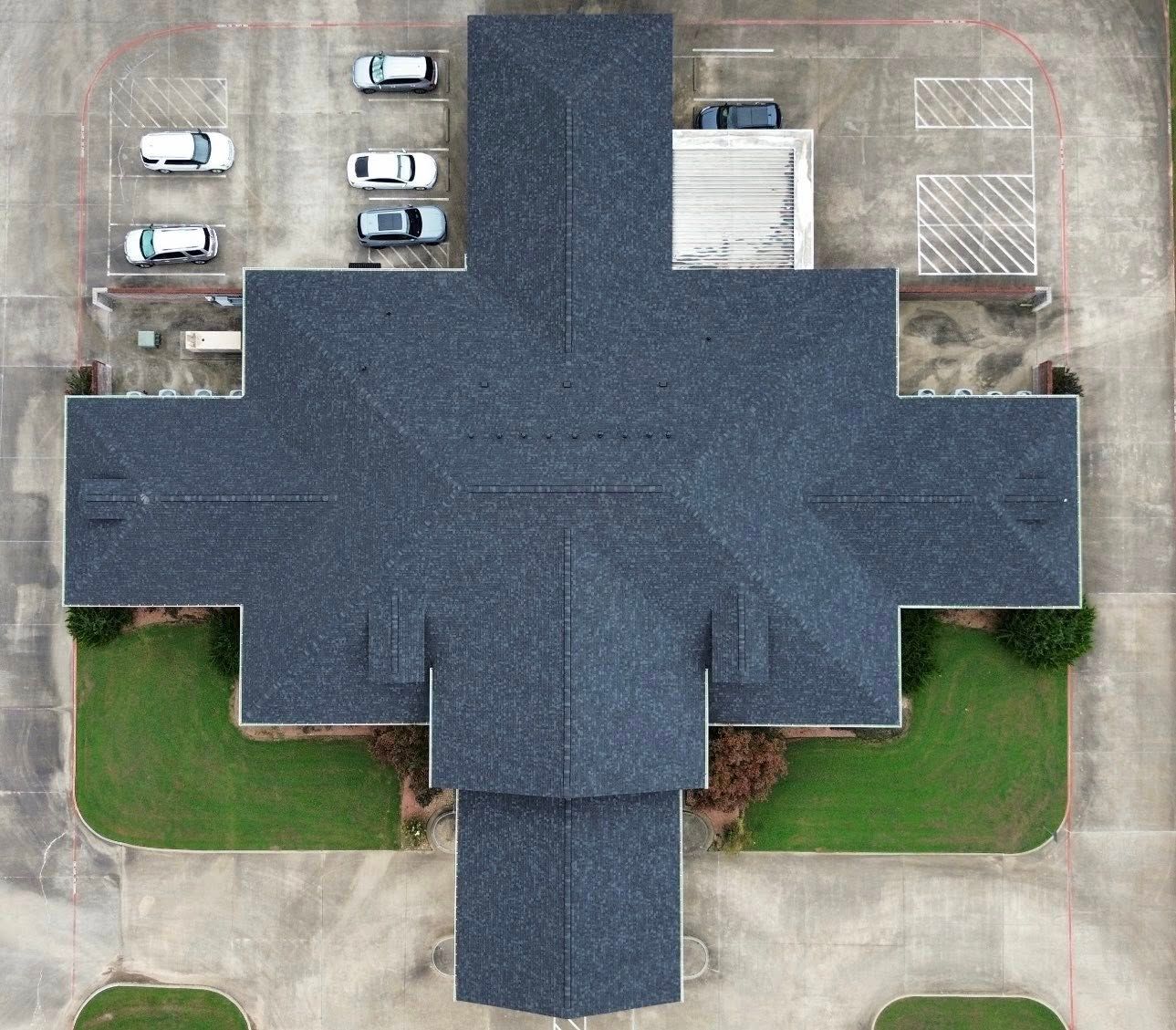 Overhead view of a building with a cross-shaped roof, parking lot, and patches of green grass.
