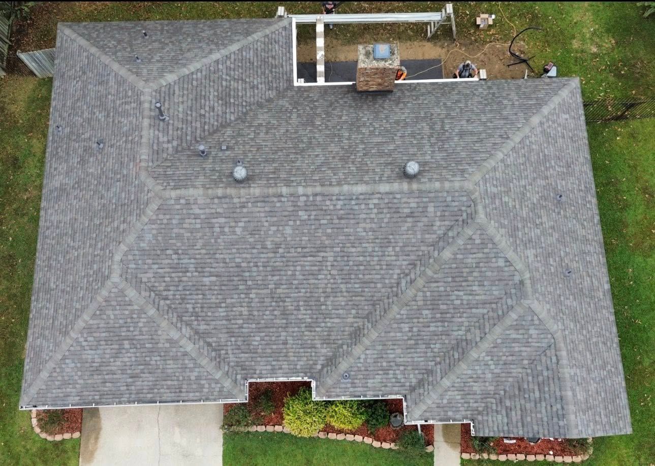 Overhead view of a gray shingled roof of a house, chimney, two vents, and lawn.