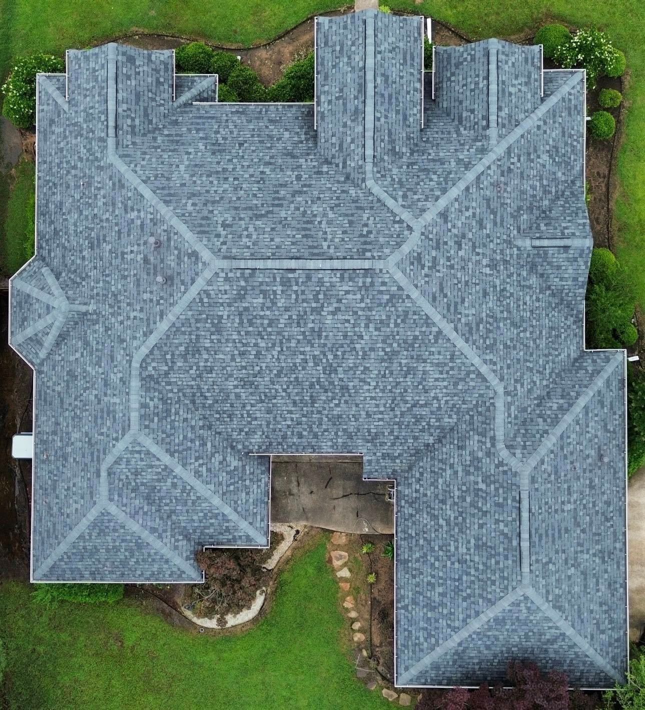 Overhead view of a gray-roofed house with a complex roof design, surrounded by green grass and trees.