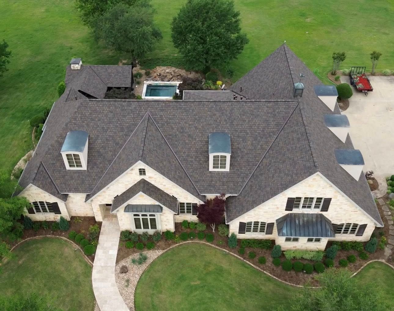 Aerial view of a large beige home with a dark gray roof, pool, and green lawn.
